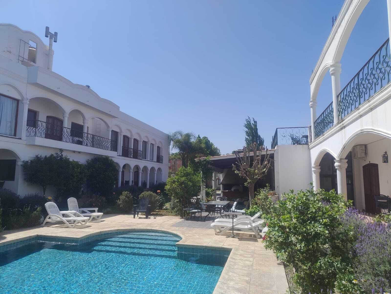 Uma área de piscina com espreguiçadeiras e uma mesa de jantar, rodeada por edifícios brancos com varandas em arco.