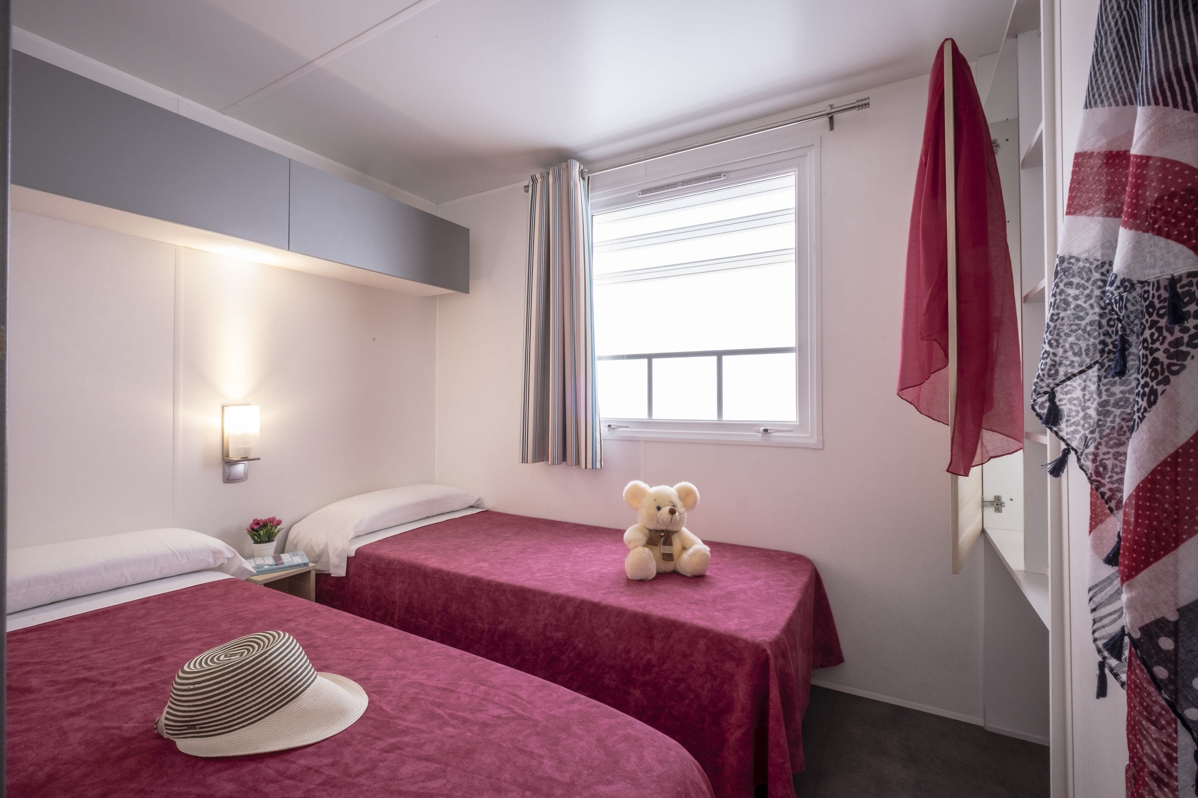 A hotel room with two beds, a teddy bear on one bed, a hat on the other, a window with curtains, and a quilt hanging on the wall.