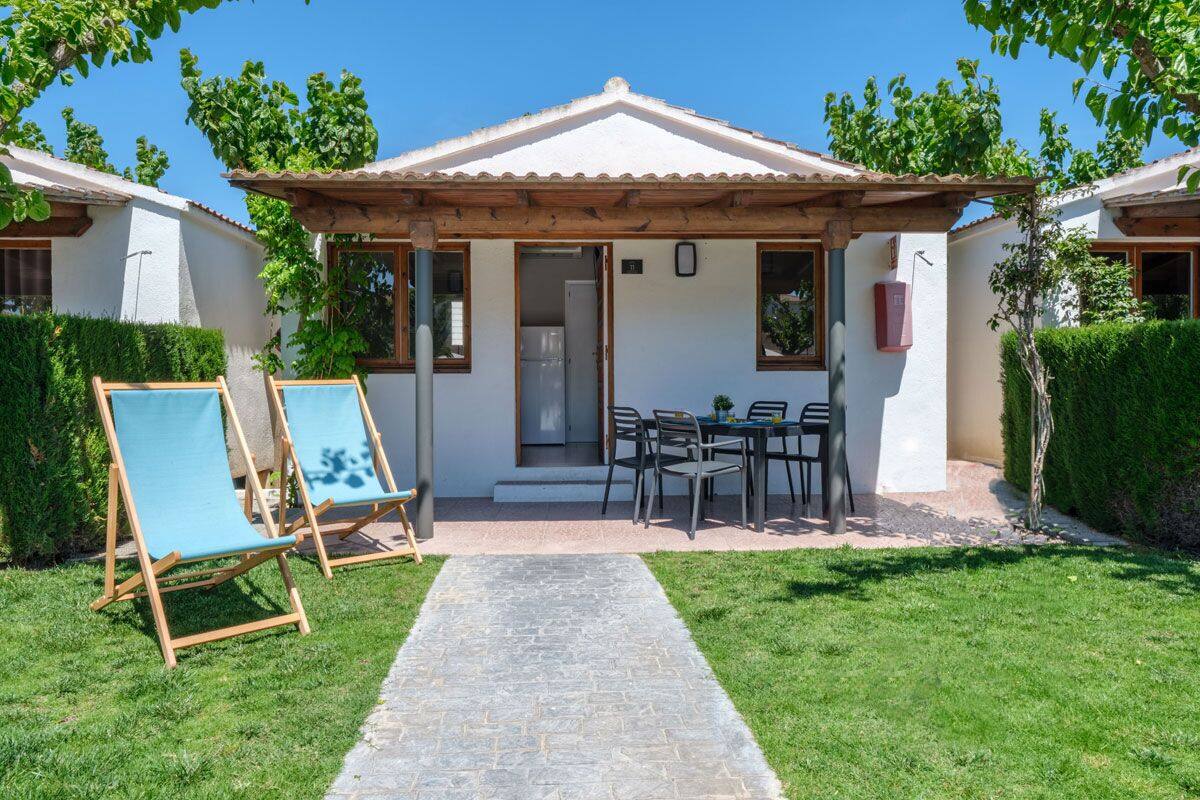 A small house with a tiled roof, a patio with a table and chairs, and a pathway leading to the entrance.
