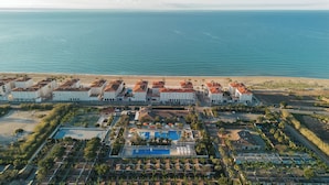 Vue aérienne d’un hôtel situé sur la côte, composé de plusieurs bâtiments, avec une piscine et une plage en arrière-plan.