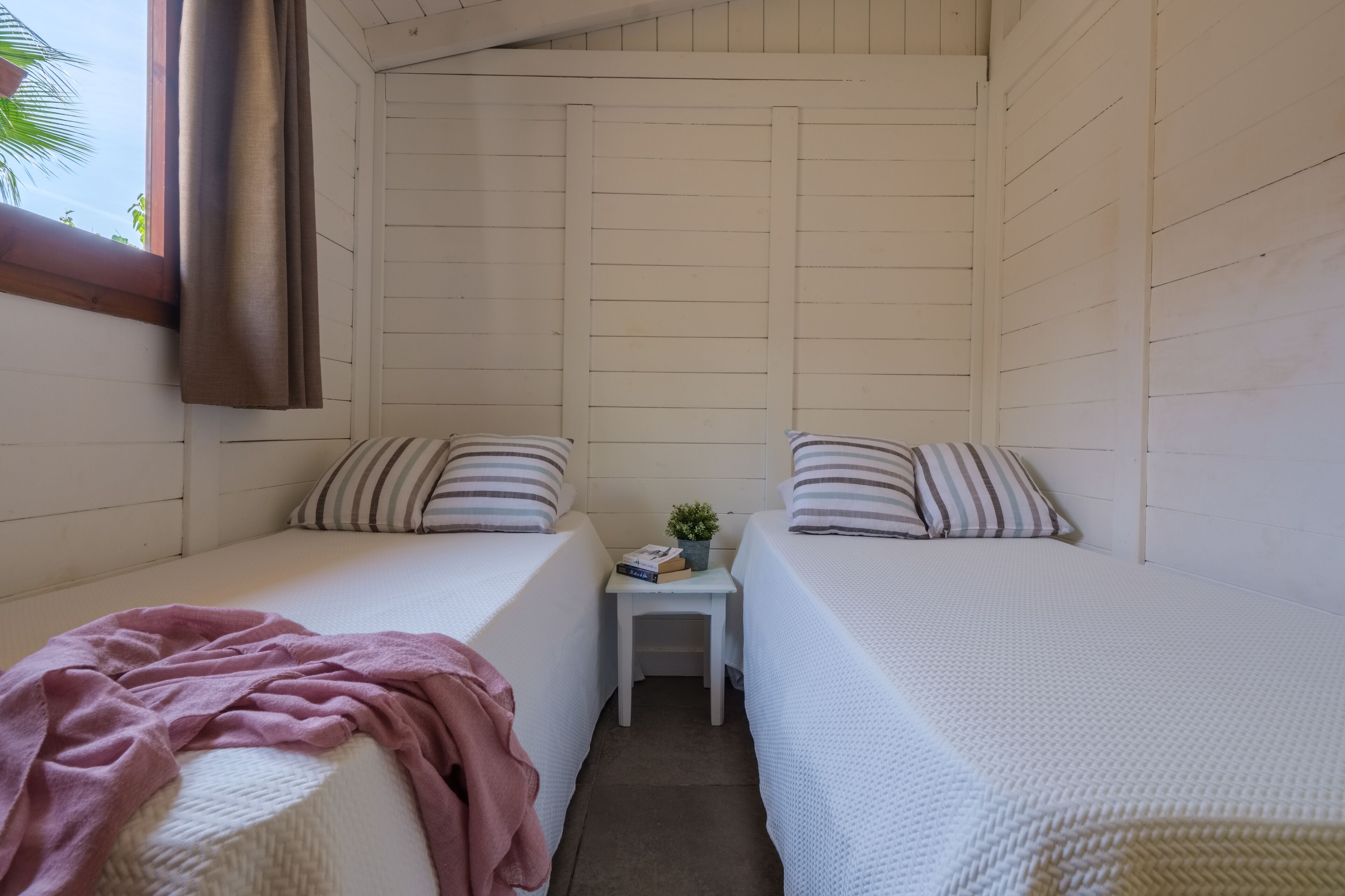 Um quarto pequeno e acolhedor com duas camas de solteiro, uma mesinha com uma planta e uma janela com cortinas.