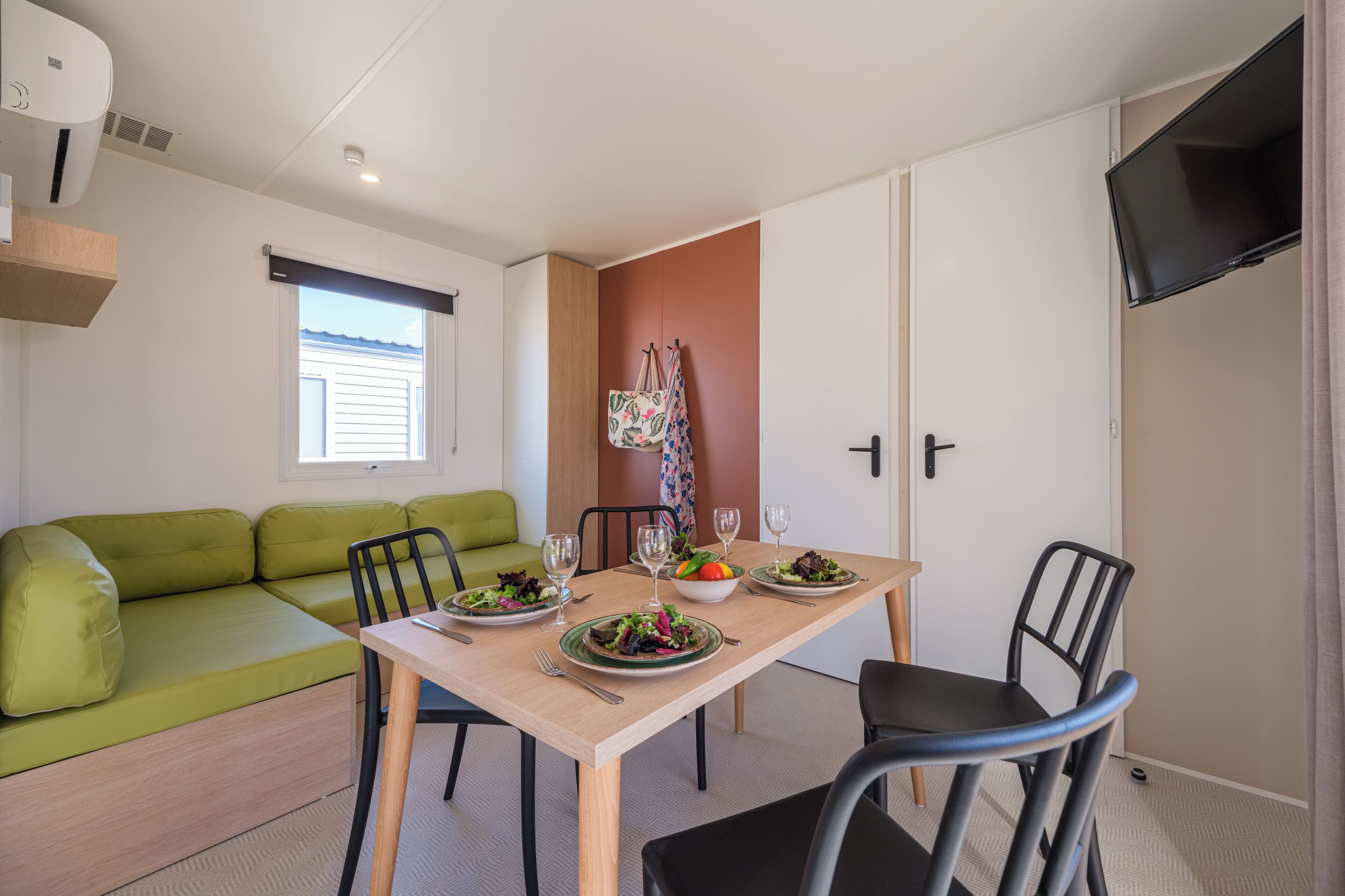 A compact living space with a dining table set for two, a green sofa, and a television mounted on the wall.
