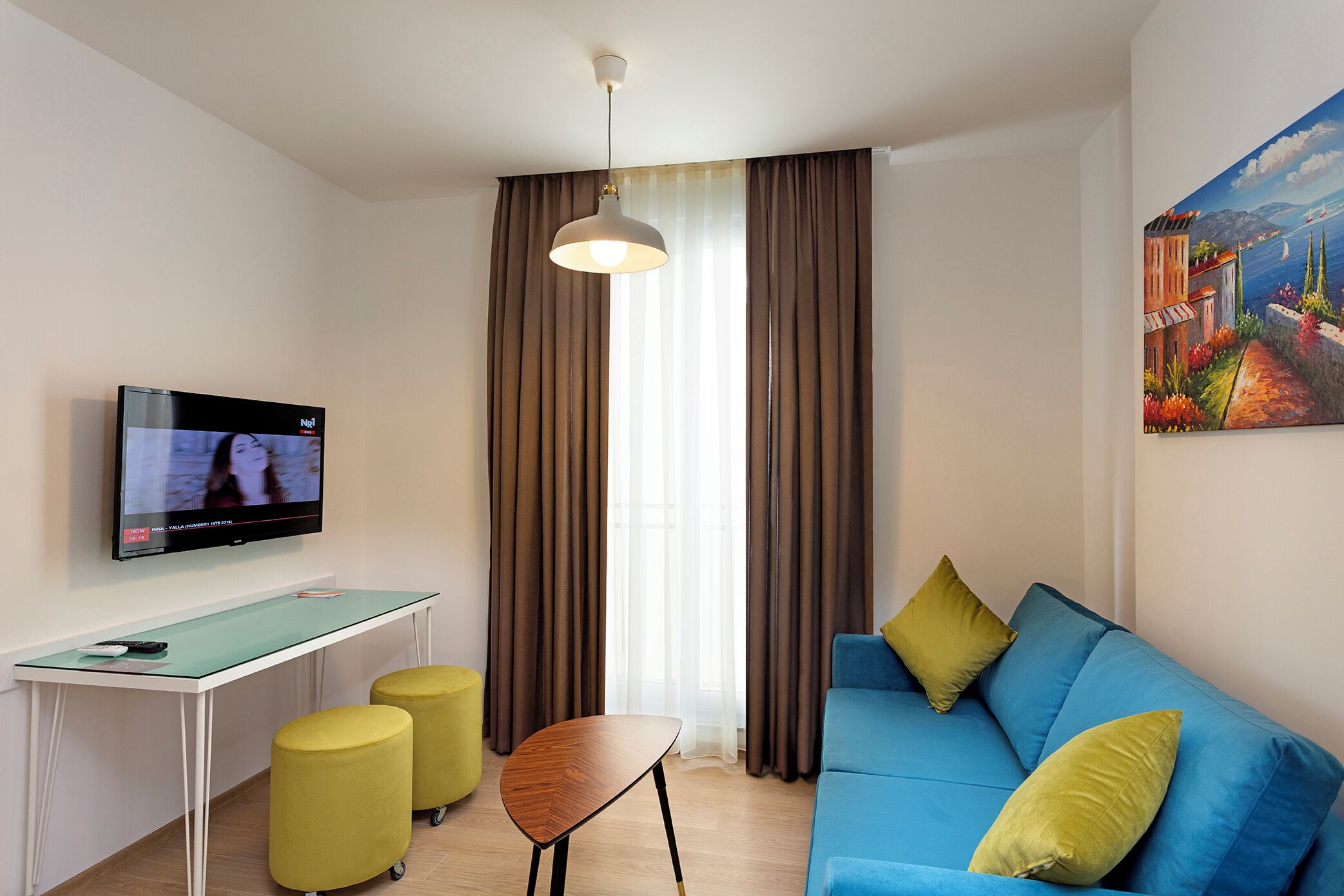 Ein modernes Wohnzimmer mit einem blauen Sofa, gelben Hocker, einem Glastisch, einem Fernseher und einem Bild an der Wand.
