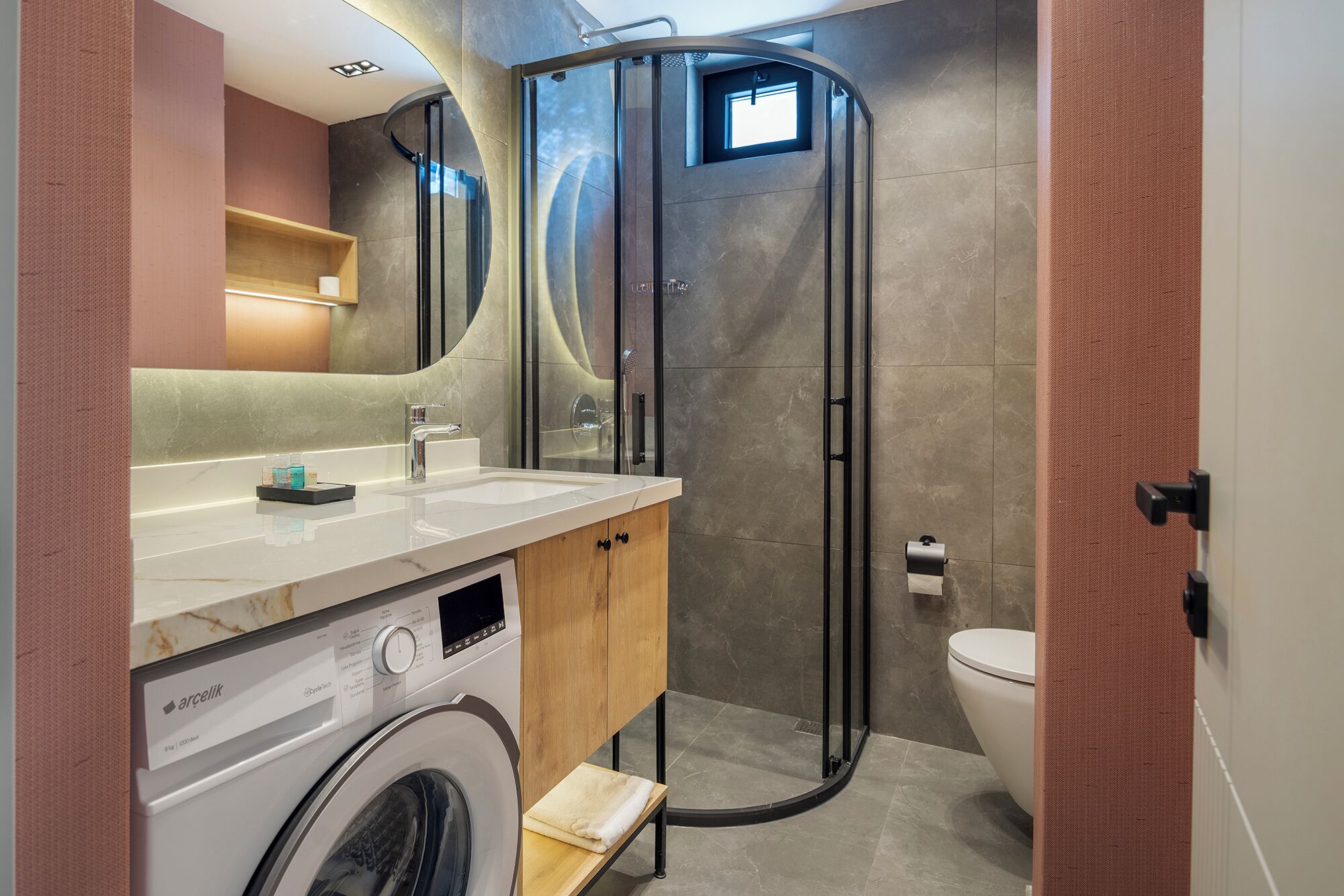 A compact bathroom with a washing machine, a toilet, a shower enclosure, and a mirror.