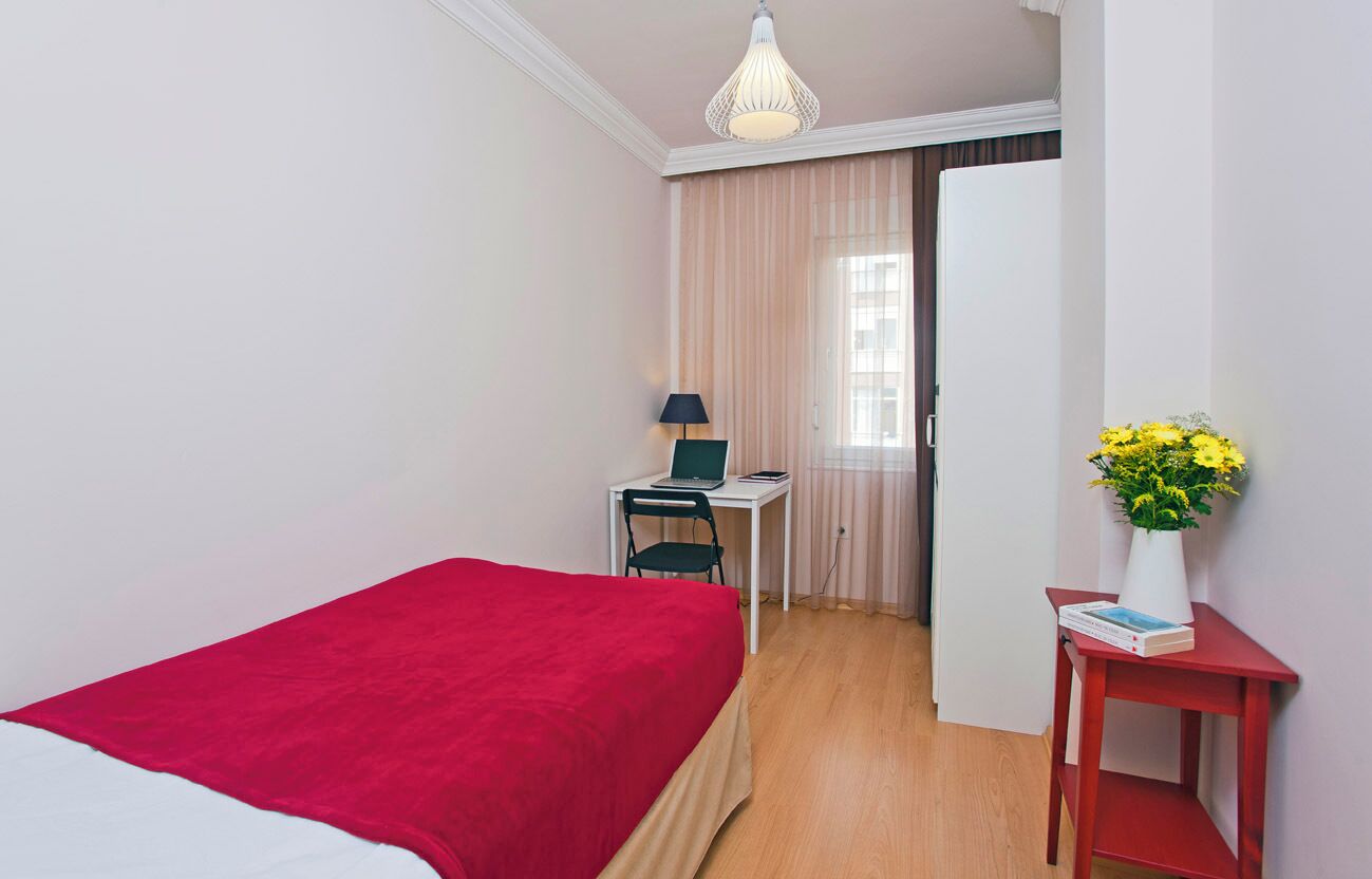 A bedroom with a bed, a desk with a chair, a lamp, a vase with flowers, and a window with curtains.