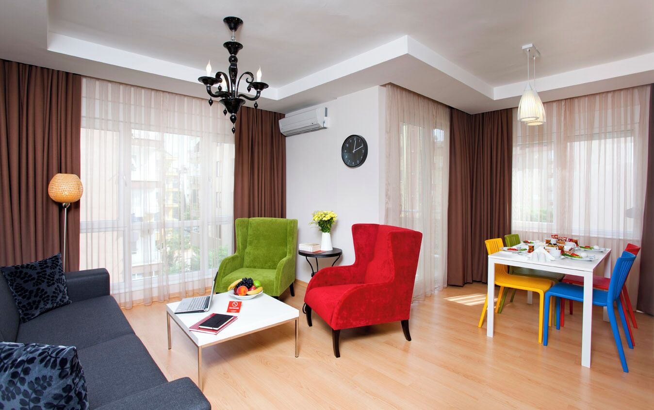 Una sala de estar moderna con sofá, dos sillones, una mesa de comedor con sillas coloridas, una araña y un reloj en la pared.