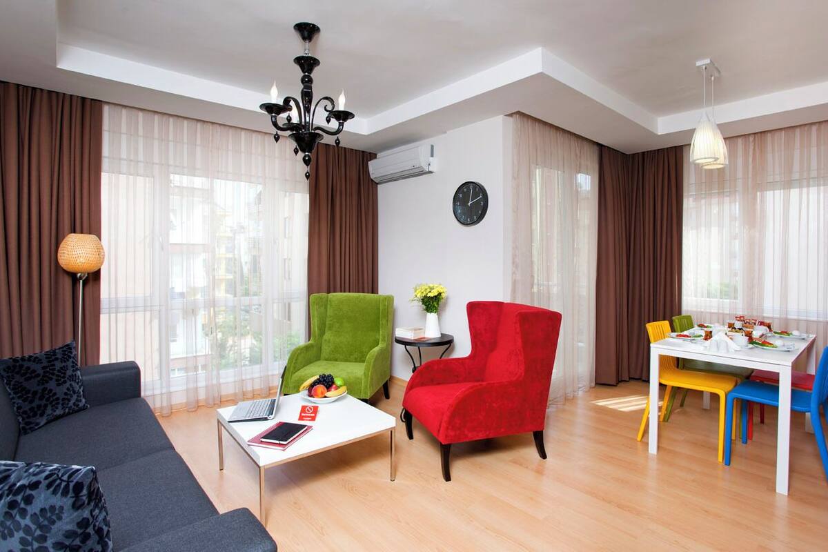 A modern living room with a sofa, two chairs, a dining table with colorful chairs, a chandelier, and a clock on the wall.
