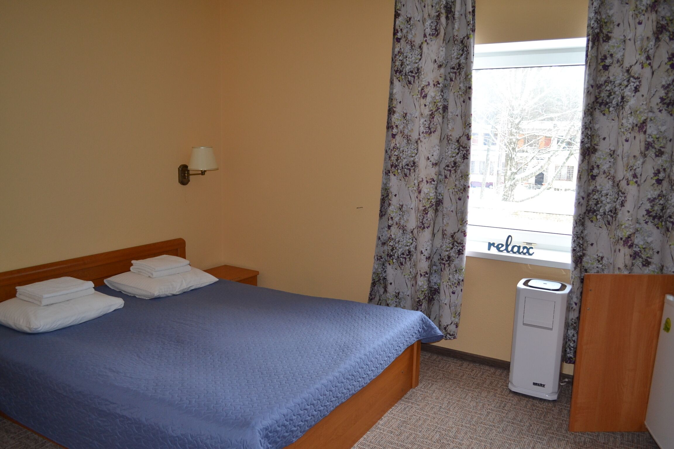 A hotel room with a bed, a wall-mounted lamp, a window with curtains, and a thermostat.