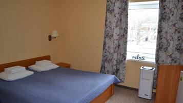A hotel room with a bed, a wall-mounted lamp, a window with curtains, and a thermostat.