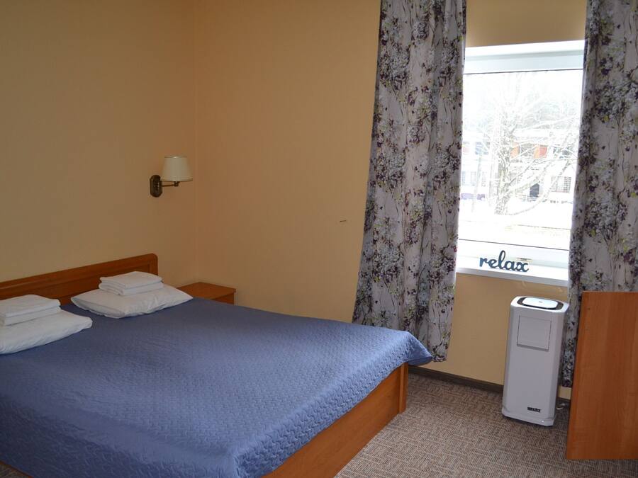 A hotel room with a bed, a wall-mounted lamp, a window with curtains, and a thermostat.