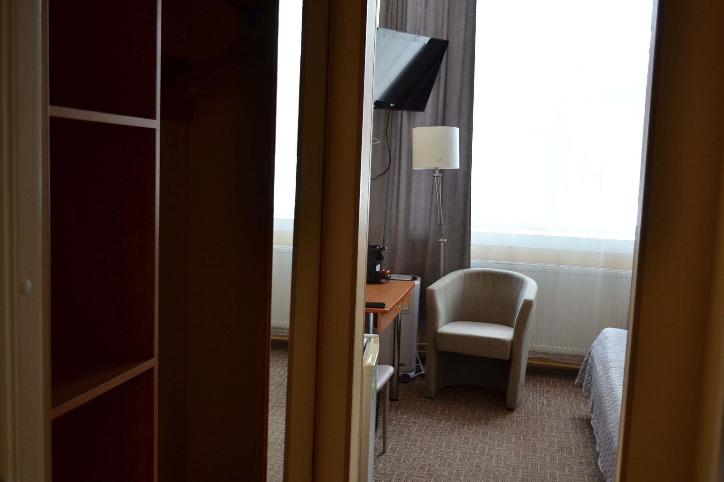 Quarto de hotel com cama, cadeira, escrivaninha com luminária e televisão na parede.