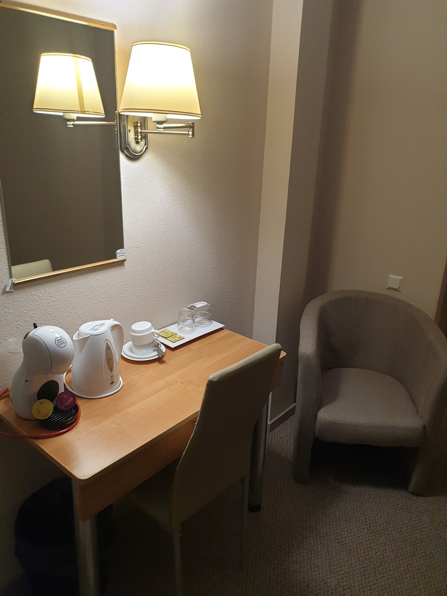 Habitación de hotel con una mesa de madera, dos sillas, un hervidor, una cafetera, un espejo y dos lámparas fijadas a la pared.