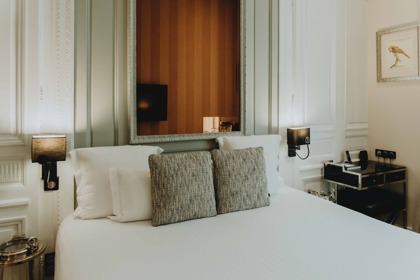 A hotel room with a large bed, two pillows, a nightstand with a lamp, a desk with a phone, and a framed picture on the wall.