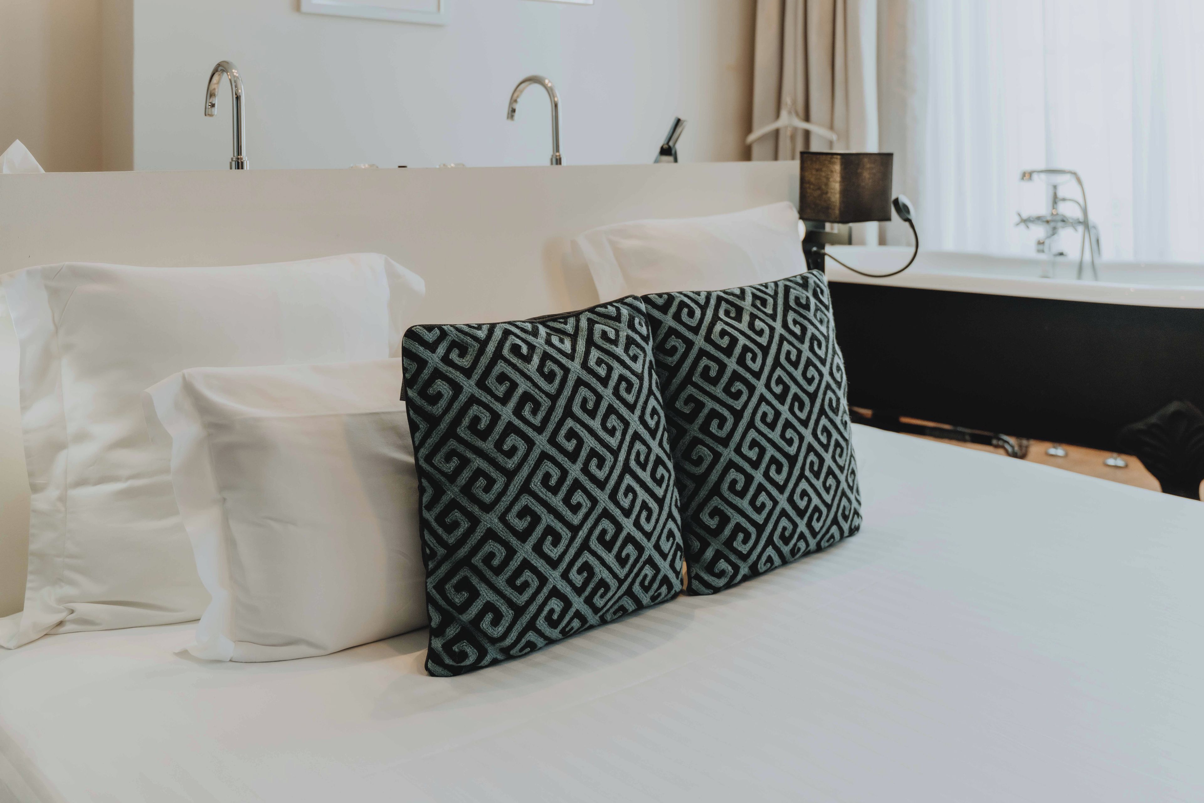 Ein Hotelzimmer mit Bett, Kissen, Nachttisch, Lampe und einem Fenster mit Vorhängen.