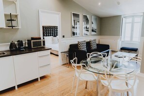 A modern kitchen with white cabinets, a dining area with a glass table and white chairs, a black sofa with patterned cushions, and a large window with artwork. - Le Boutique Hotel & Spa (Bordeaux)