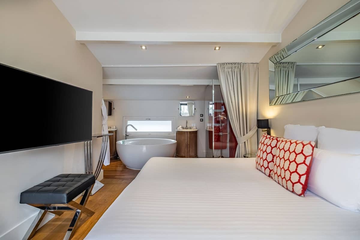 A modern hotel room with a large bed, a bathtub, a flat-screen TV, and a wooden floor.