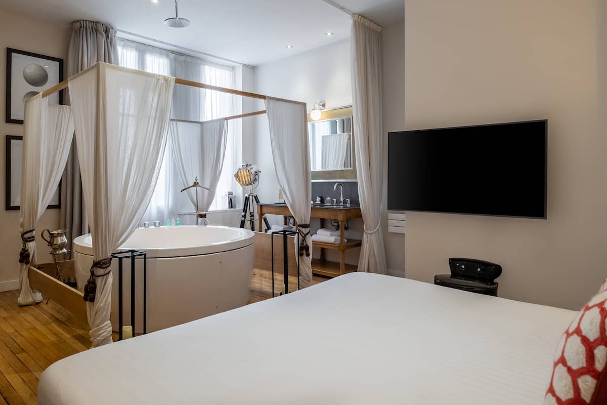 A modern bedroom with a large bed, a freestanding bathtub, a vanity with a mirror, and a flat-screen TV mounted on the wall.