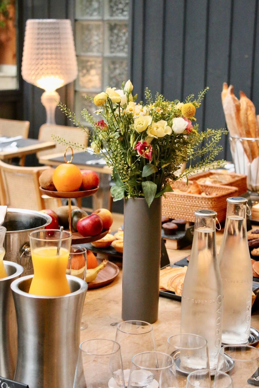 A breakfast setting with a vase of flowers, fruit bowl, juice pitchers, and bread.