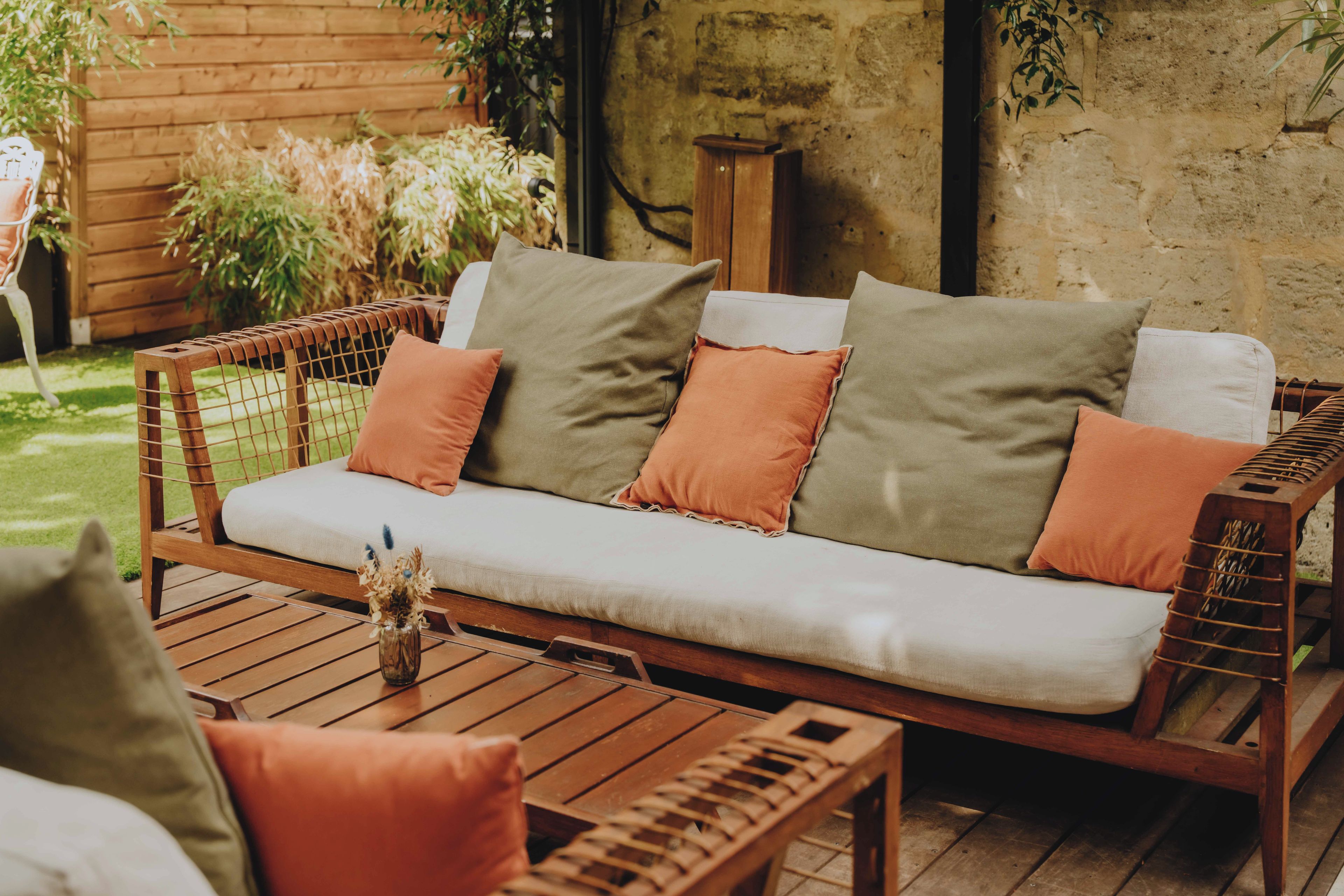 A wooden outdoor seating area with a cushioned bench and a table, surrounded by plants and a rustic wall.