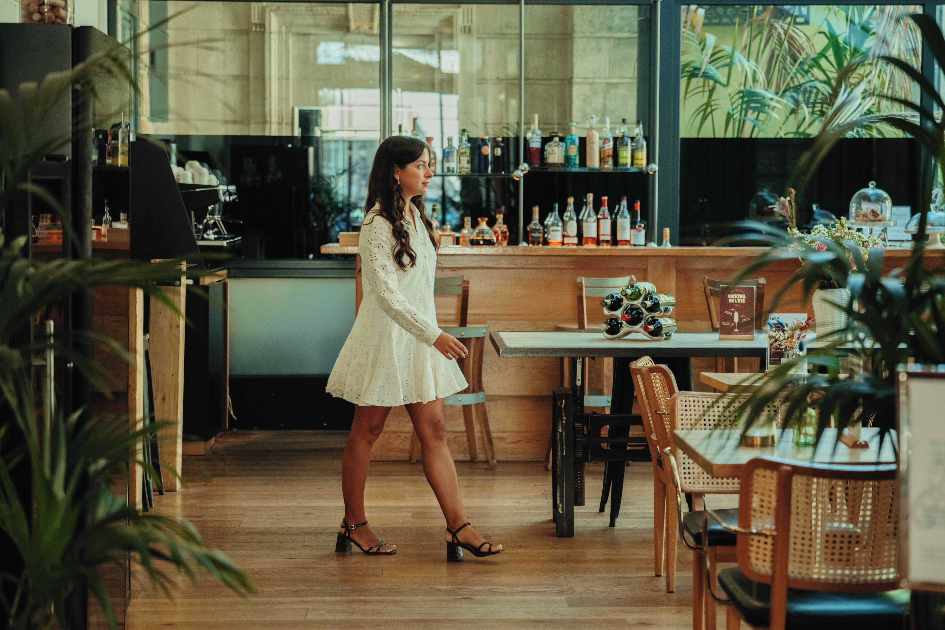 A woman in a white dress standing in a bar area with wooden flooring, bar stools, and a variety of bottles on shelves.