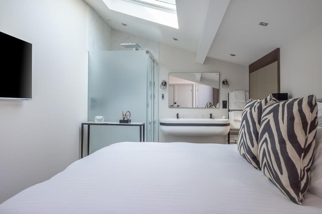 A modern hotel room with a large bed, a glass shower enclosure, a sink, and a mirror.