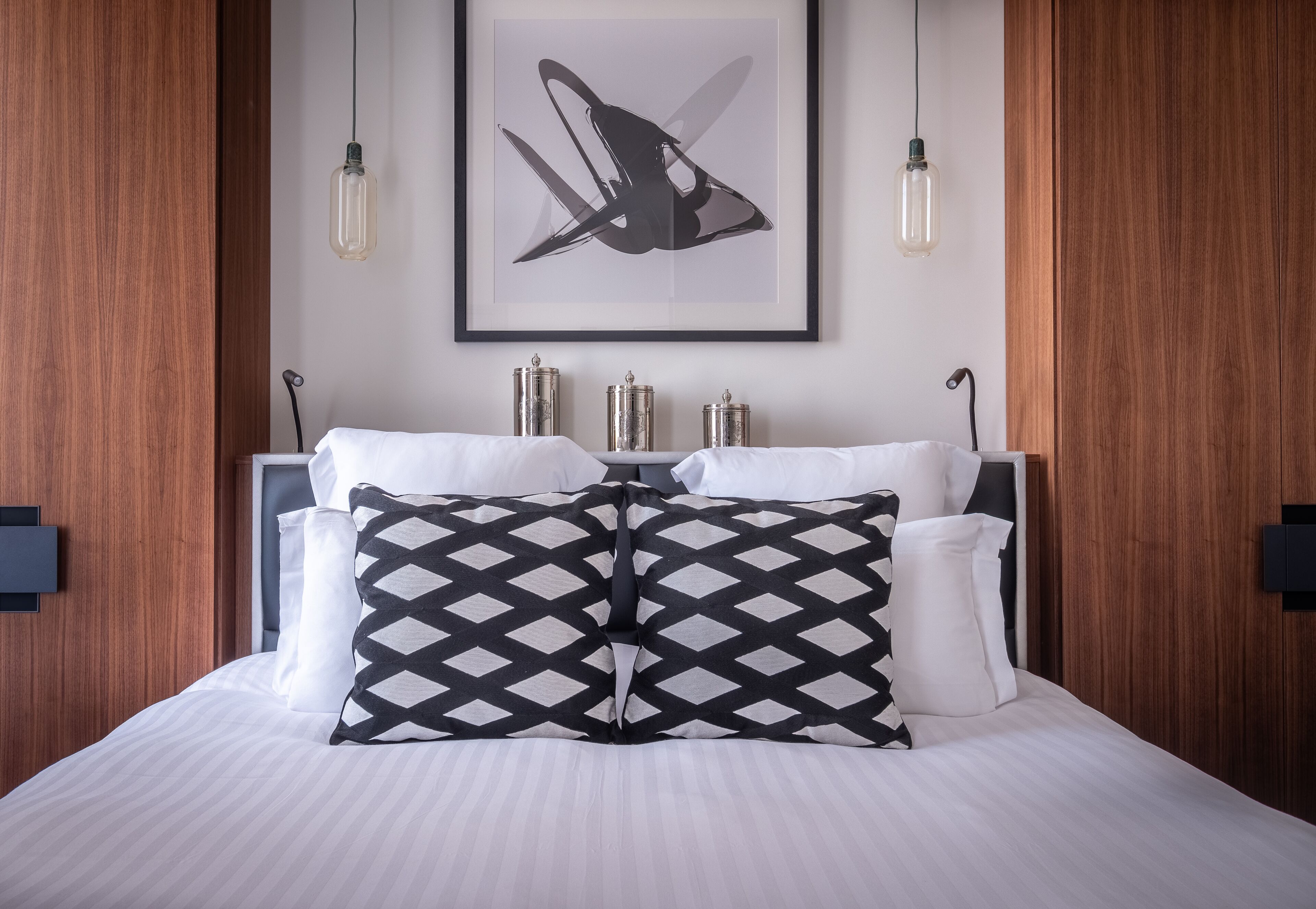 Una cama bien hecha con sábanas blancas y dos almohadas estampadas, una obra de arte abstracta enmarcada encima, y dos mesitas de noche con recipientes metálicos.