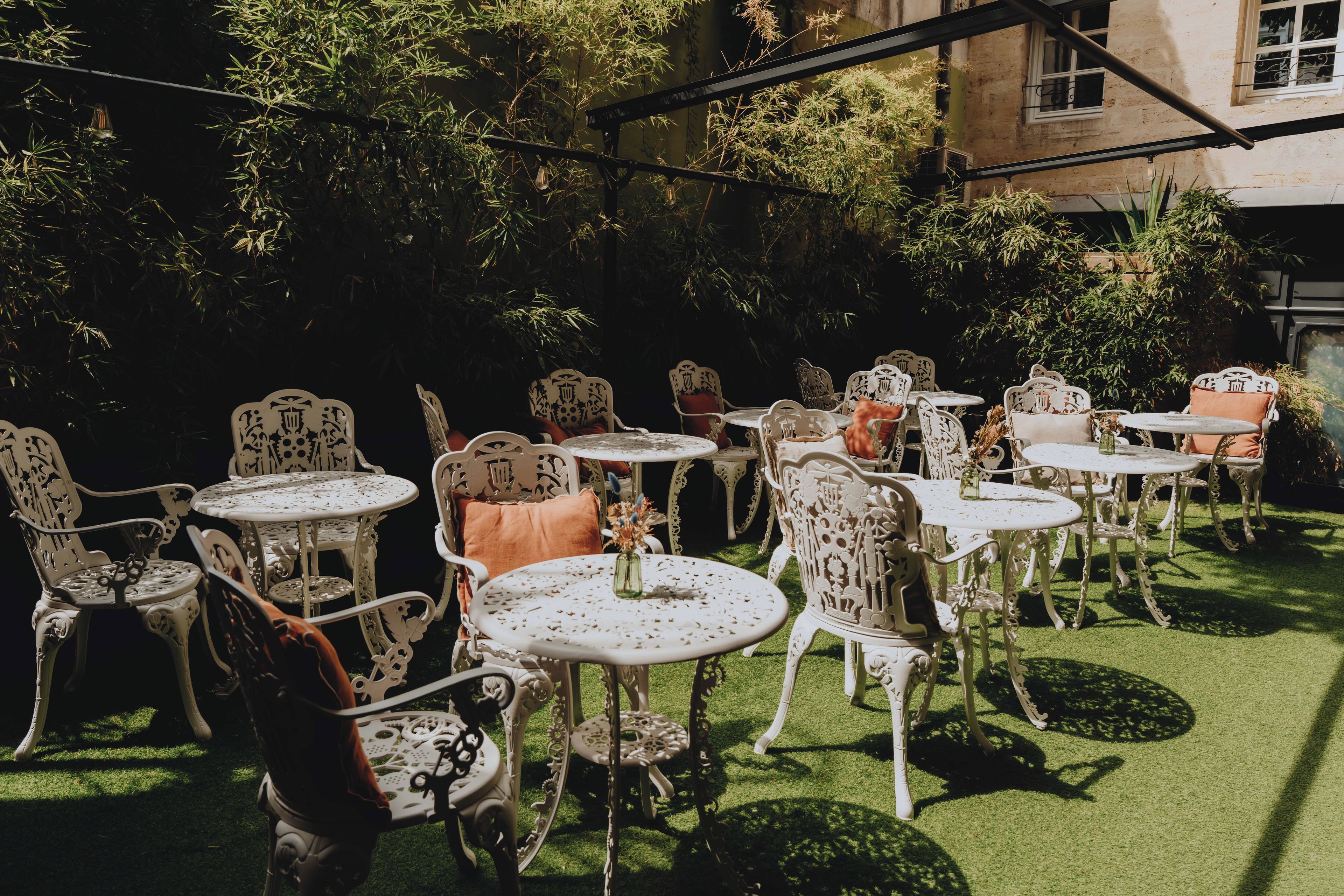 An outdoor seating area with ornate metal chairs and tables, surrounded by lush greenery and a building in the background.