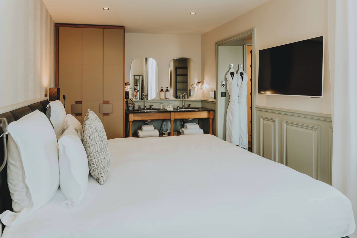 A hotel room with a large bed, a double sink, a mirror, and a flat-screen TV mounted on the wall.