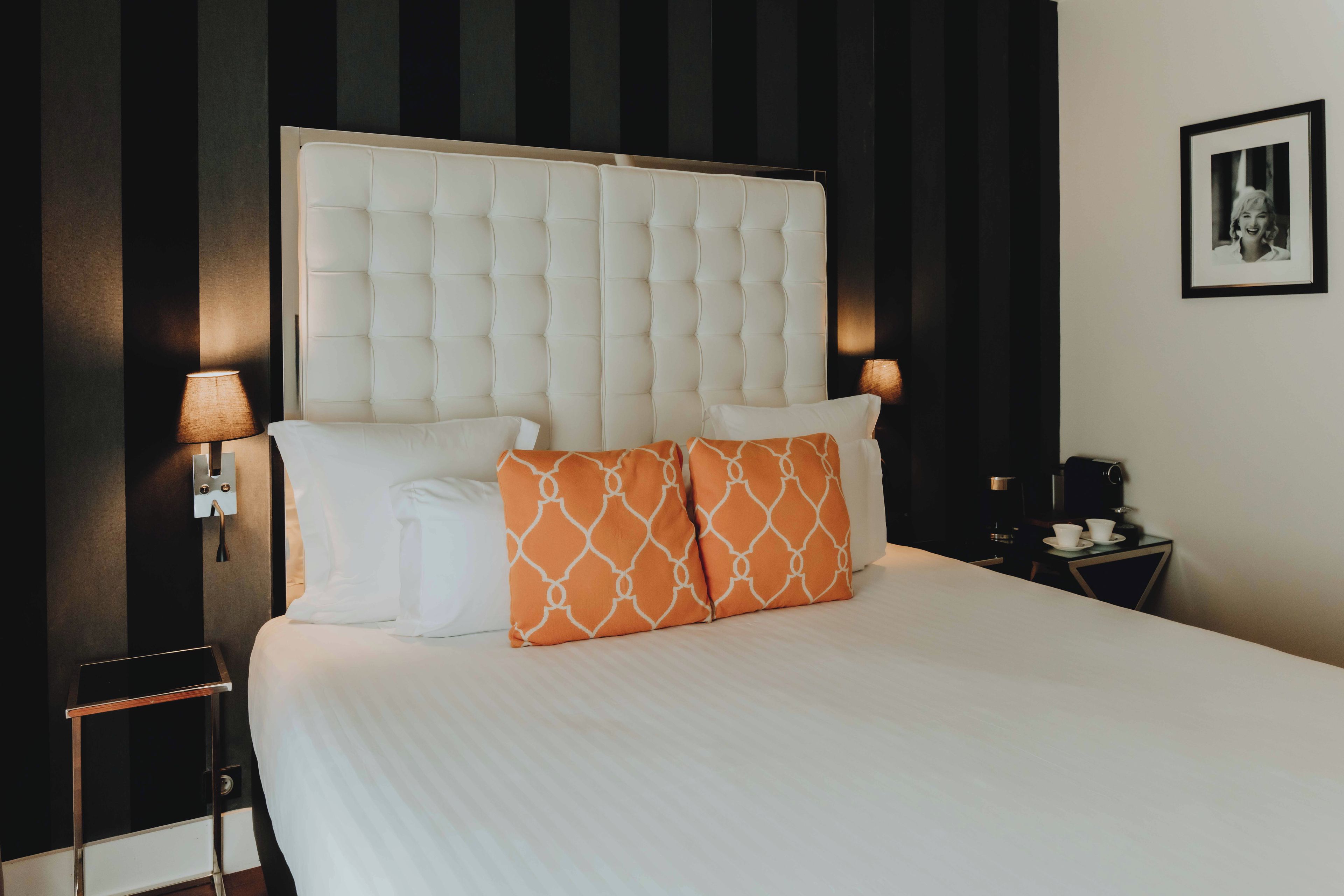 A neatly made bed with a tufted headboard, white bedding, and two pillows, one white and one orange with a geometric pattern.