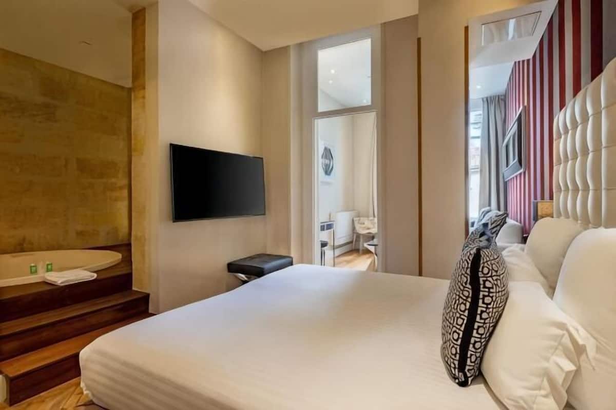A hotel room with a bed, a TV mounted on the wall, a bathroom with a sink and mirror, and a view of the outside.