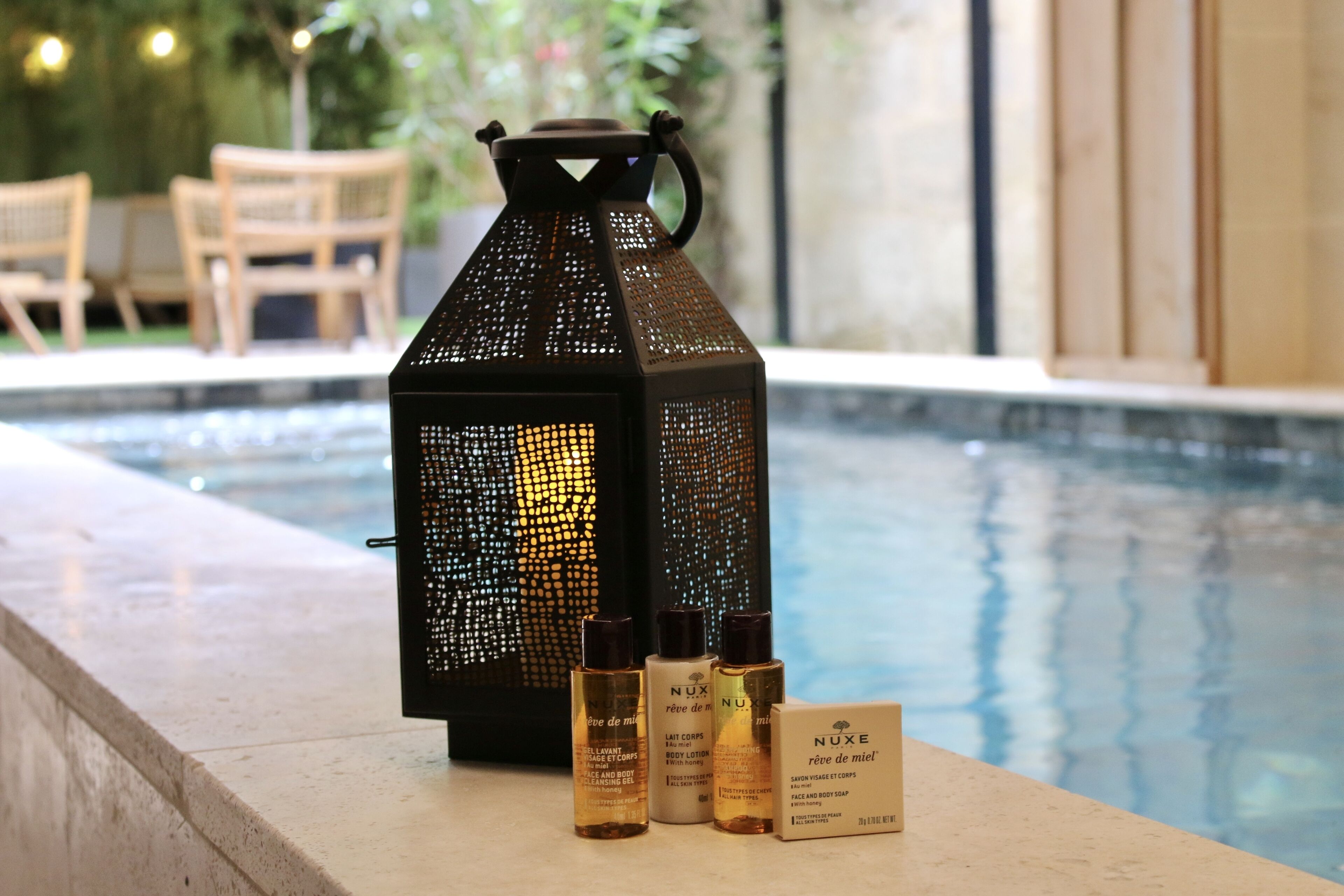 Escena junto a la piscina con un farol decorativo, tres botellas de productos para el cuidado corporal Nuxe y una silla de madera al fondo.
