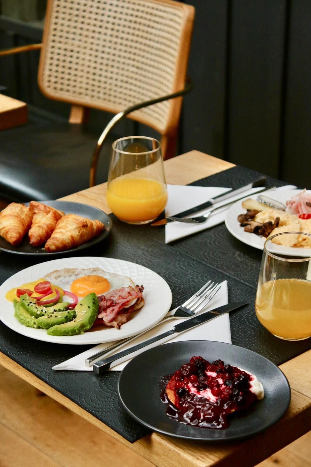 Une table de petit-déjeuner avec des croissants, des œufs, de l’avocat, du bacon et des fruits rouges sur une assiette.