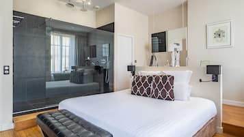 Ein modernes Hotelzimmer mit einem großen Bett, einer separaten Duschkabine, einem Schminktisch mit Spiegel und einem gerahmten Bild an der Wand.