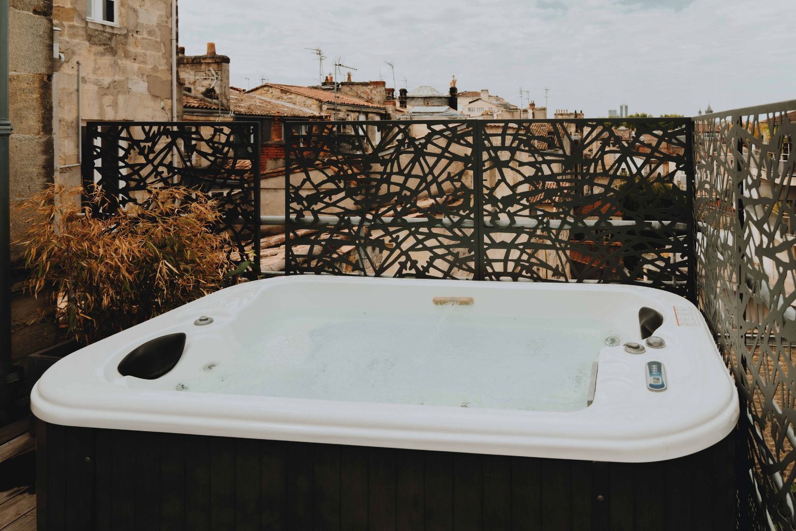 Un jacuzzi sur un balcon, agrémenté d’une rampe en métal décorative, avec vue sur les bâtiments en arrière-plan.