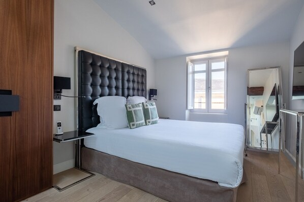 A modern bedroom with a tufted headboard, a large window, a mirror, and a wooden wardrobe.
