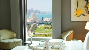 Quarto de hotel com uma janela ampla que oferece vista para o horizonte urbano e um corpo d'água. Há duas poltronas e uma cama com colcha florida.