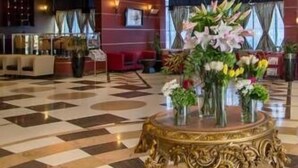 Um hall de entrada com um lustre grande, uma mesa redonda dourada com arranjos florais e um chão quadriculado.