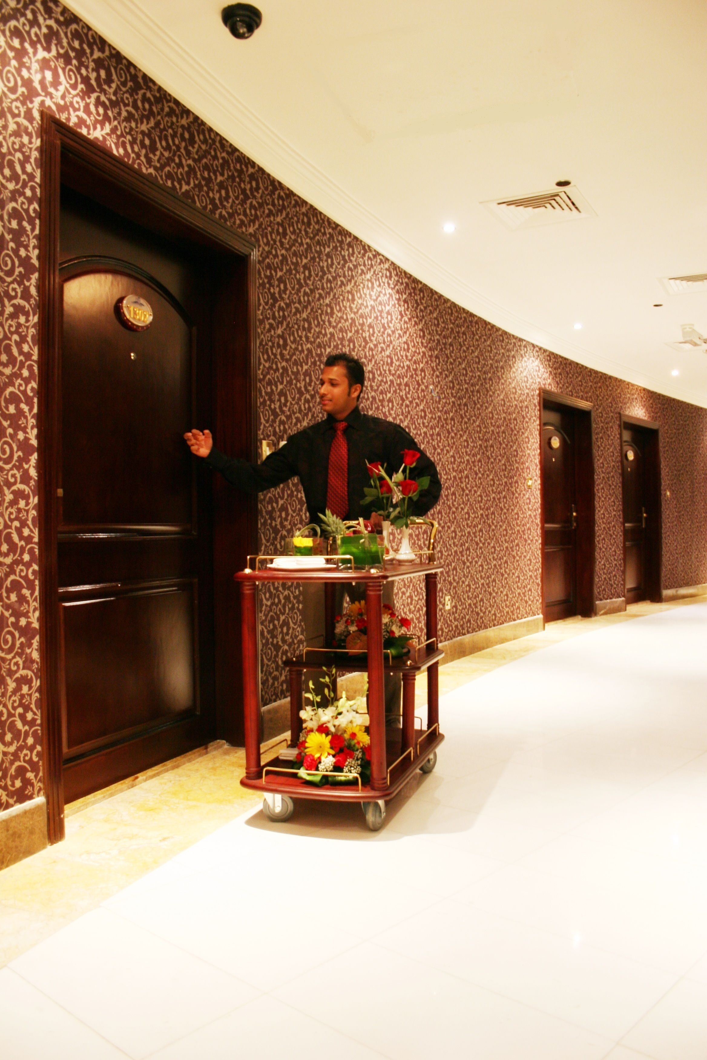 A man in a black suit and red tie standing next to a cart with flowers and a bottle.