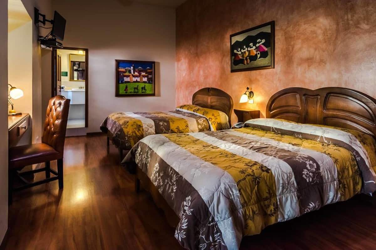 Une chambre d’hôtel avec un plancher en bois, deux lits avec une literie à motifs floraux, une tête de lit en bois, un bureau avec une chaise et un tableau accroché au mur.