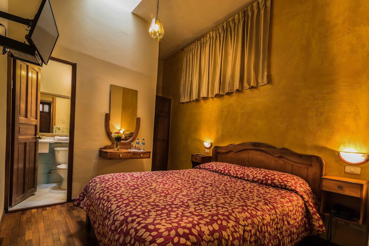 Une chambre d’hôtel avec un grand lit, une tête de lit en bois, une table de chevet avec une lampe, un miroir et une vue sur la salle de bain.