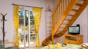 A room with a wooden staircase, a television on a wooden stand, a table with yellow mugs, a chair, and a sofa. - Madame Vacances Les Mas de St Hilaire (Saint-Hilaire-de-Riez)