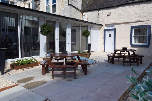 A courtyard with a wooden table and benches, surrounded by a white building with large windows and a door. - The Crown Hotel (Newton Stewart)