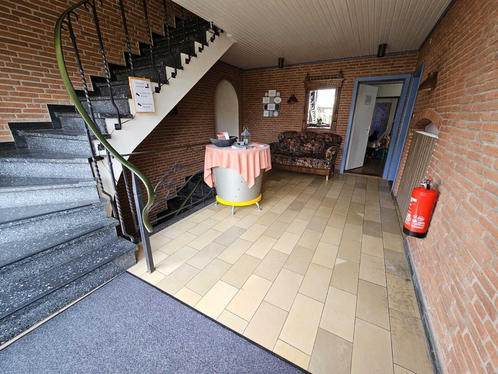 Eine Treppe führt zu einem Raum mit Fliesenboden, einem Tisch mit einer rosa Tischdecke, einem Sofa und einem an der Wand befestigten Feuerlöscher.