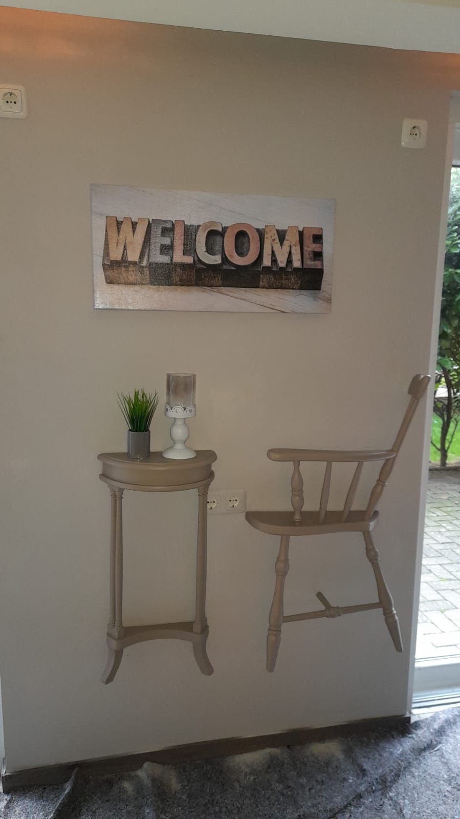 A wall with a 'WELCOME' sign, a small table with a plant, and a wooden chair.