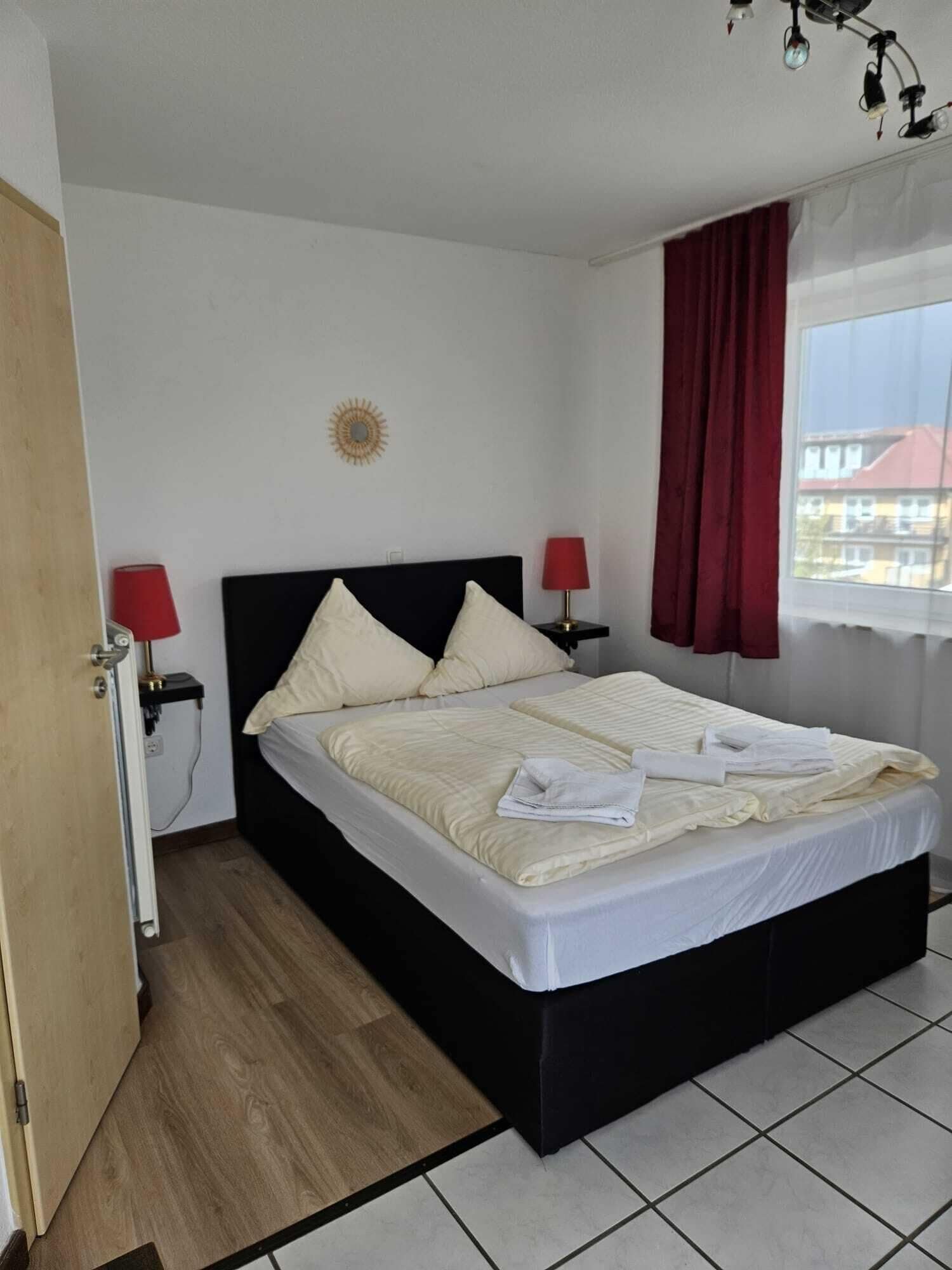 A hotel room with a large bed, two bedside lamps, a window with curtains, and a view of buildings outside.