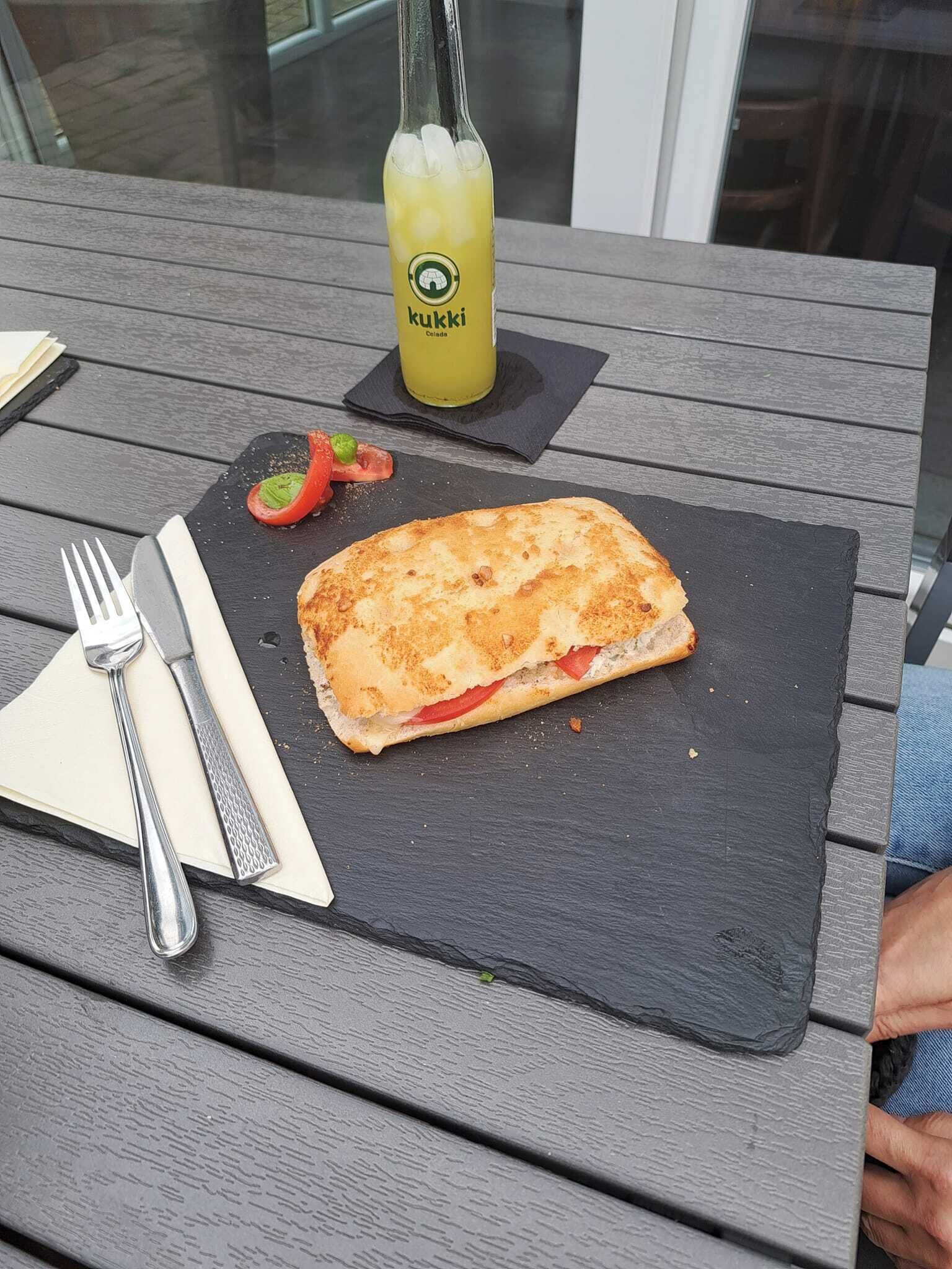 Um sanduíche em uma tábua de ardósia com uma garrafa de suco e um garfo sobre uma mesa de madeira.