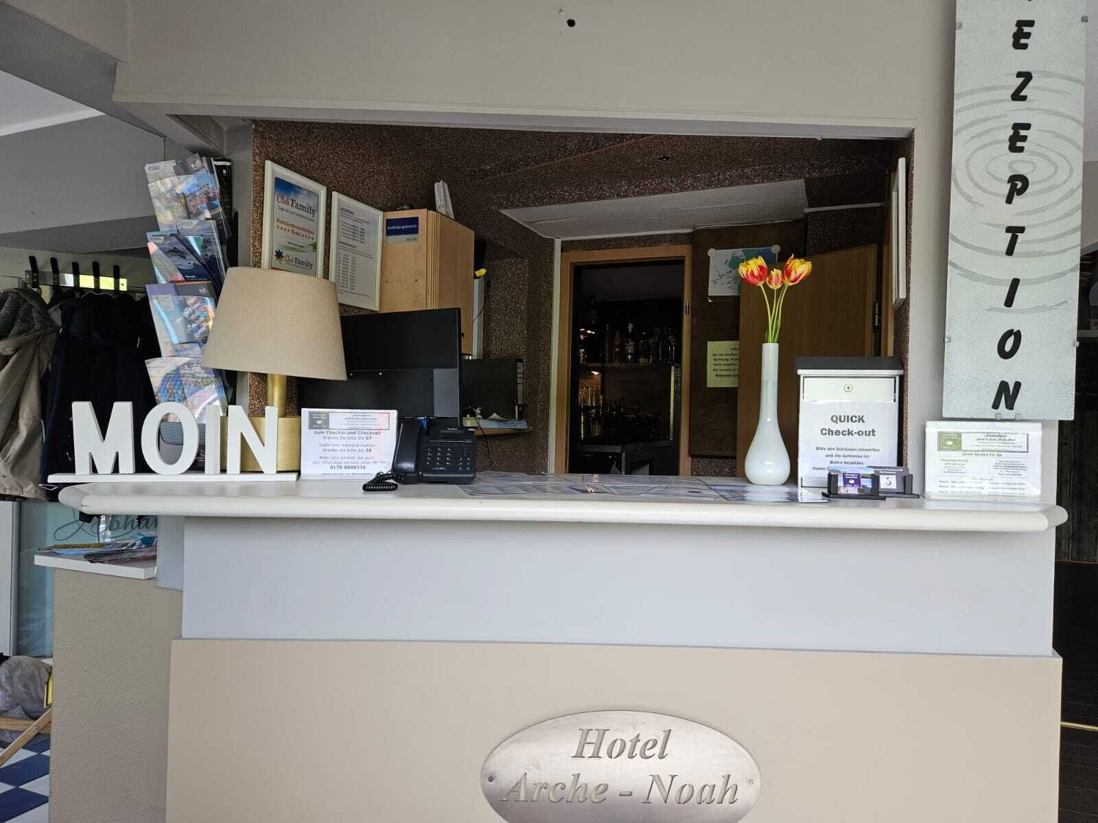 A hotel reception desk with a 'MOIN' sign, a 'Hotel Arche - Noah' plaque, a 'QUICK Check-out' sign, and a vase with flowers.