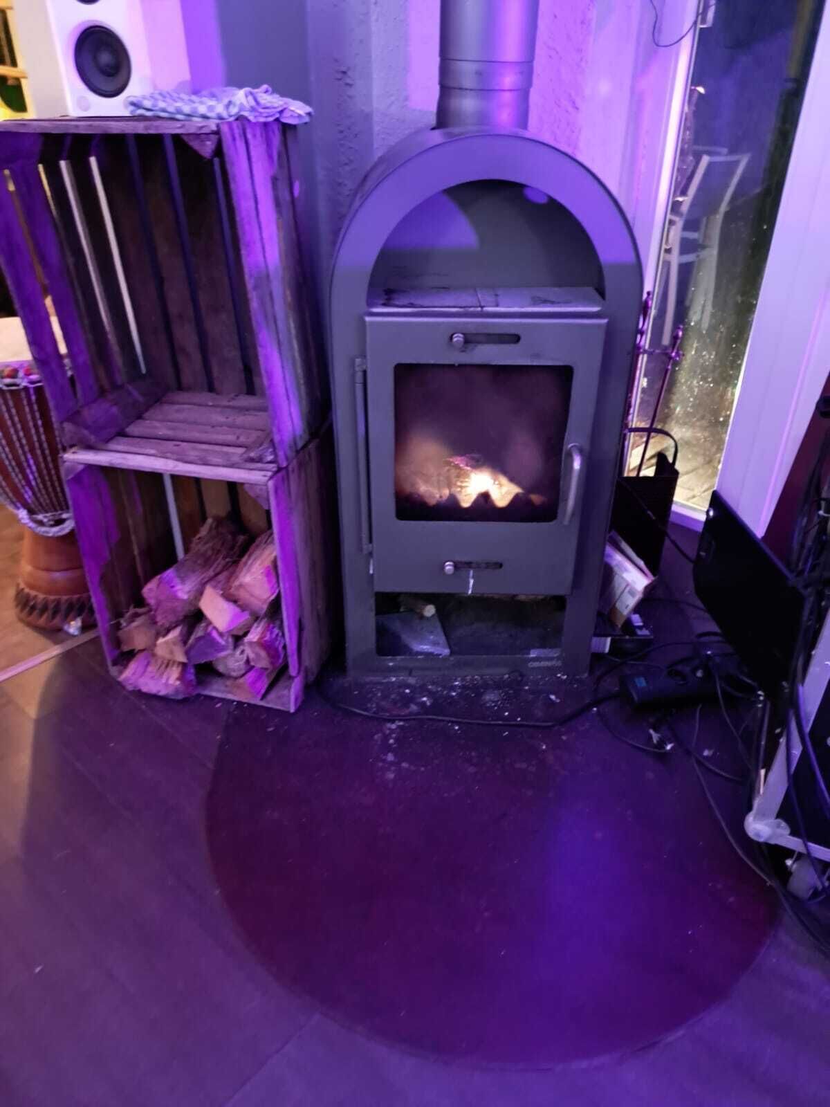 A wood-burning stove with a fire burning inside, a wooden shelf with firewood, and a window with a view of the outside.