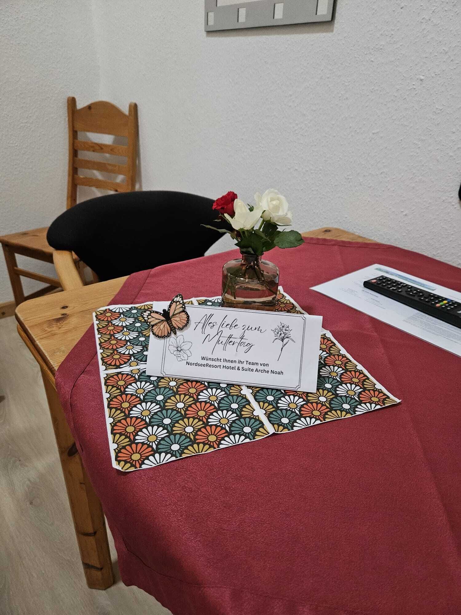 A table with a red tablecloth, a vase with white and red flowers, a remote control, and a card with text in German.