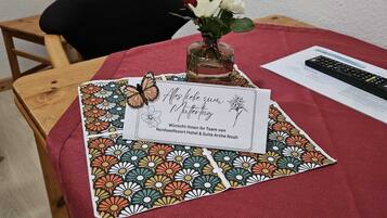 A table with a red tablecloth, a vase with white and red flowers, a remote control, and a card with text in German.